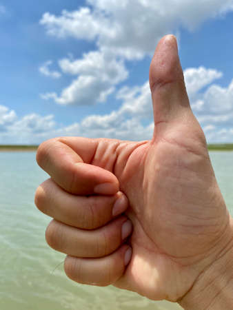 Like sign, Thumb up, Good Job for complementing of Asian male hand with sky water and grass on sunny day with sky and cloud as a background in Lopburi Thailand. Close up.の写真素材