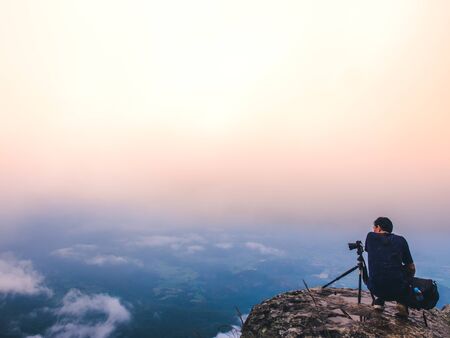 Nature photographer takes photos with camera on tripod at cliff. Dreamy foggy landscape. Morning...