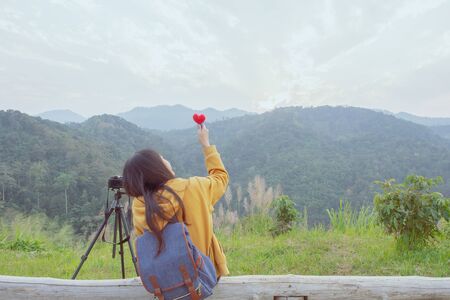 backside of hipster girl with backpack, enjoying sit see to valley landscape view on peak mountains in tropical rainforest. show the heart shape. solo traveling concept.の写真素材