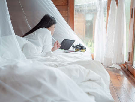 Young Asian woman freelance working on notebook and tablet remotely while rest in bedroom of resort...