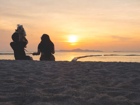 Pets love concept from silhouette of couple and dog sitting together on the beach at sunsetの写真素材