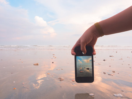 Holding smartphone and taking photo of the beach at sunset from low angle.の写真素材