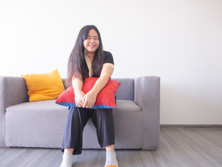 Young woman sits on sofa hugging cushion, smiling and relaxing. Simple pleasures at home concept.の写真素材