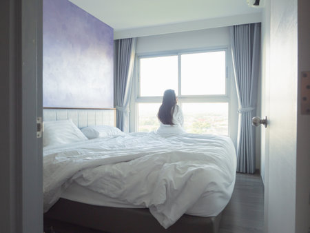 Young woman relaxing in bed with white bedding, smiling in soft morning light by the windowの写真素材