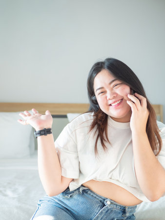 Asian woman smiling while talking on the phone in cozy bedroom. Bright natural light creates warm and relaxed morning atmosphere at home.の写真素材