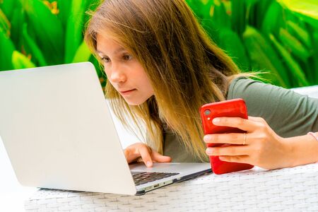 Blond teenager girl using laptop while holding a red smartphoneの写真素材