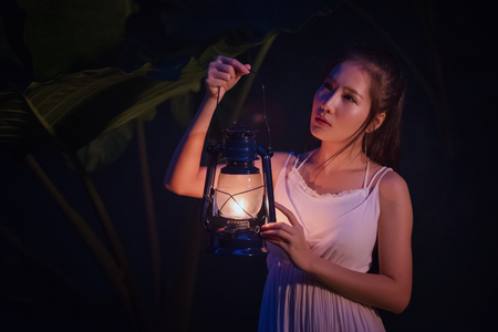 Woman with Vintage Lantern Outside at Night - Cosplay girl in nutral  costume holding a lamp, Young woman in white long dress walking in night woodの写真素材