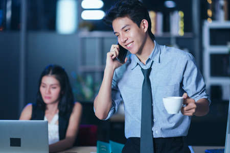 Businesspeople in the office at night working late, coffee and  calling phone working overtime together over a laptop at a desk in an office in the late eveningの写真素材