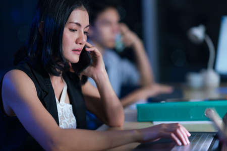 Businesspeople in the office at night working late, coffee and  calling phone working overtime together over a laptop at a desk in an office in the late eveningの写真素材