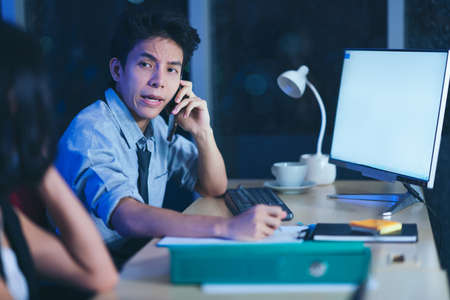 Businesspeople in the office at night working late, coffee and  calling phone working overtime together over a laptop at a desk in an office in the late eveningの写真素材