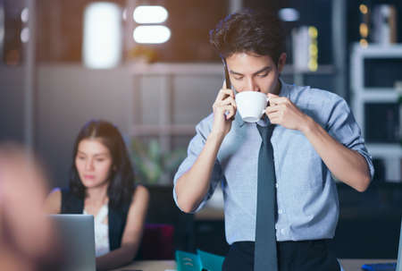 Businesspeople in the office at night working late, coffee and  calling phone working overtime together over a laptop at a desk in an office in the late eveningの写真素材