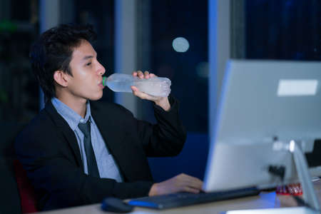 Young asian handsome business man drinking water after work hard in office at night, businessmen relax after working, business success conceptの写真素材