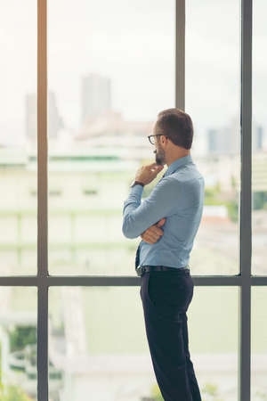 Adult male businessman, teacher, mentor looks out the large window of his office. Work in the office. Development of a new business projectの写真素材