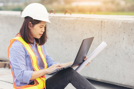 Beautiful Asian female engineer in white safety hard hat using laptop computer notebook doing job at...