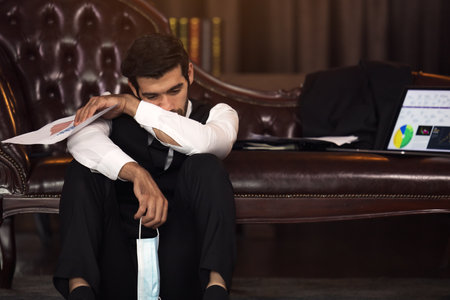 Depresses businessman with closed eyes holding hands on head and paper with charts in officeの写真素材