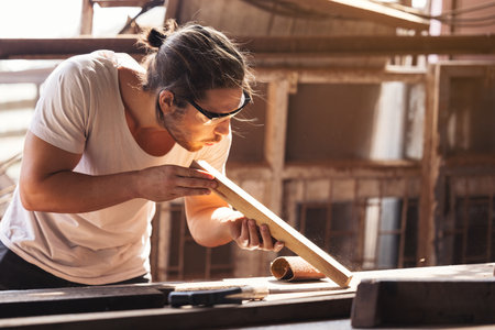 A young male carpenter builder working overall equals a wooden bar with a milling machine in the...