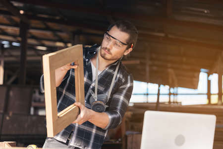 A young male carpenter builder working overall equals a wooden bar with a milling machine in the...