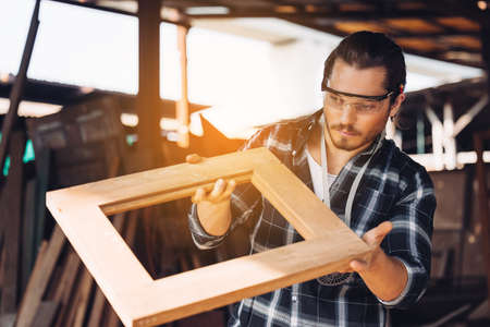 A young male carpenter builder working overall equals a wooden bar with a milling machine in the workshop, in the background woodenの写真素材