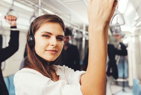 Beautiful young woman passenger standing with headphones and while shirt in the modern subway trainの写真素材