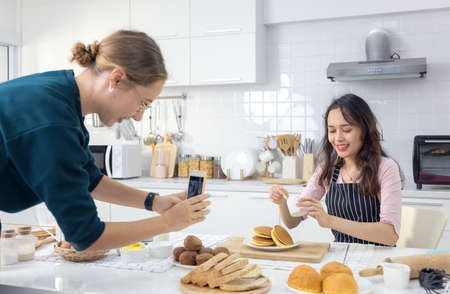 Beautiful woman pastry chef kneading bread dough on the worktop, as another female snaps a snapshot of her doing so.の写真素材
