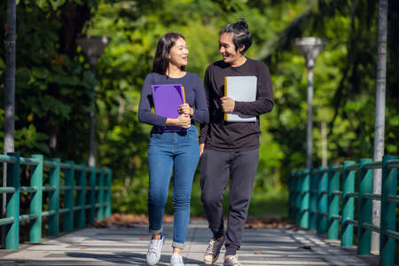 A young college student pair on their way to class. They are taking a walk around the university campus while reading a book. Autumn is a beautiful season.の写真素材