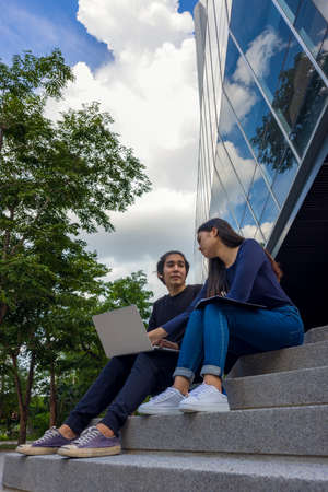 Two happy teenage students studying homework with a copybook sitting on stairs outside.