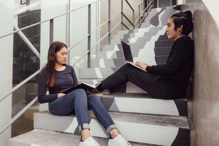 Two happy teenage students studying homework with a copybook sitting on stairs outside.