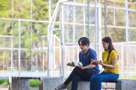 On a campus, a couple of students are studying together, and a teenager sits on a seat beside a sports court with a book.の写真素材