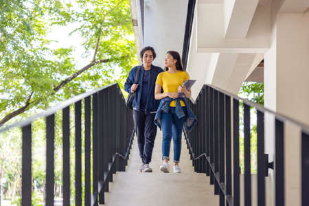 Two Asian university students walking and conversing to class in a magnificent campus building's...