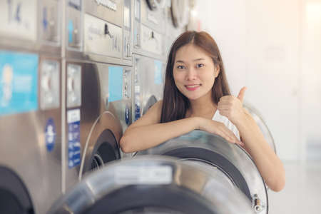 In the self-service laundry with dryer machines in the backdrop, a young woman enjoys clean ironed garments.の写真素材