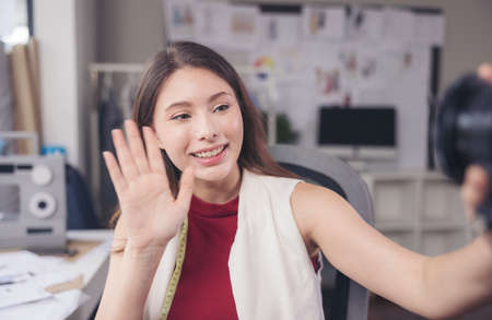 Waving her hand, a fashion designer sits at her workstation in her studio, looks at the camera online present, and communicates through video call.の写真素材