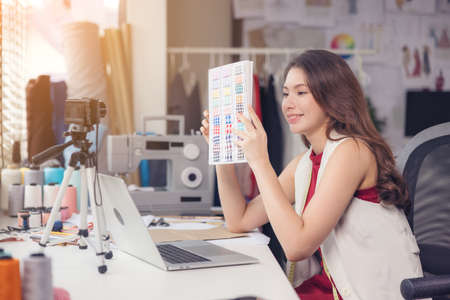 A lovely Asian woman fashion designer sits at her workstation in her studio, stares at the webcam,...