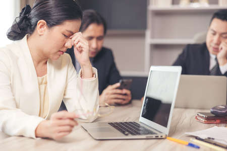 A stressed woman sits at her desk in the office. After a long period of laptop use, a tired businesswoman removes her spectacles, suffering from eye strain and dry eyes syndrome.の写真素材