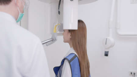 On a panoramic x-ray machine, the dentist aligns the bite of a woman. In a dental clinic, a woman dressed in a lead apron stands still as a panoramic X-ray machine circles her head.の写真素材