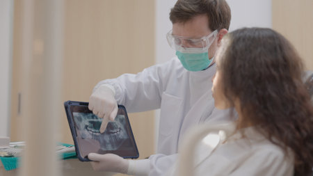 At a modern dental clinic, a dentist with a face mask discusses test results with a woman, and a Caucasian male and female dentist with a face mask use a tablet to advise a smiling Asian woman patient on how to treat her teeth.の写真素材