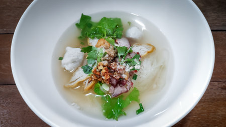 Thai style clear small noodles soup with grilled pork and fish balls in white bowl on Wooden table...