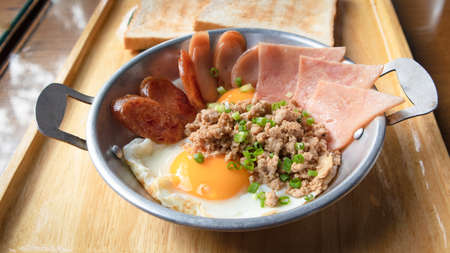 Fried eggs in a small pan with ham, minced pork, spring onion and chinese sausages on wood tray. Popular Thai Breakfast called Kai Kata.の写真素材
