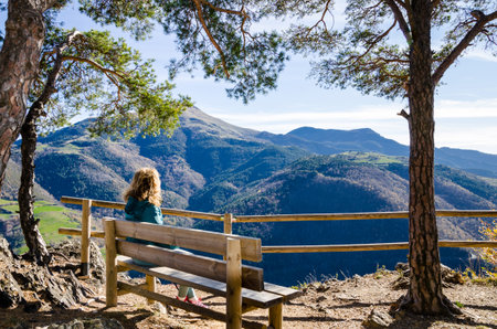 Curly-haired blonde woman, sitting on a bench, looking at the fantastic views of the Ribes mountains, in the Ripolles region, Gironaの写真素材