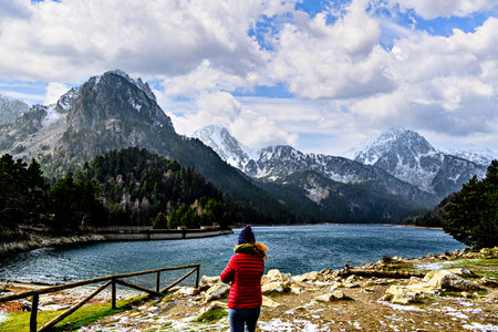 Woman in red coat at Sant Maurici Lake with snowy mountains, in Aigües Tortes National Park, in the...