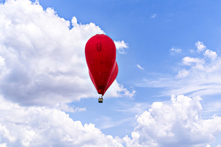 Red heart-shaped hot air balloon flying over a blue sky with white cloudsの写真素材