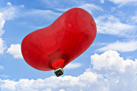 Red heart-shaped hot air balloon flying over a blue sky with white cloudsの写真素材