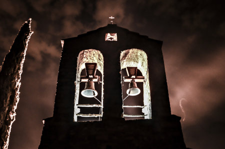 Silhouette of a church with bells under an illuminated dark and gray skyの写真素材