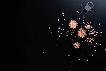 Black measuring spoons with Himalayan salt on black background.