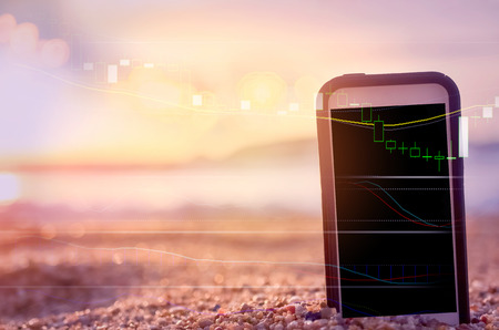 Business economic technology and freedom trader working concept. Close up smart phone on sand sunset beach double expose graph money stock currency bokeh background. Vintage tone filter color styleの写真素材