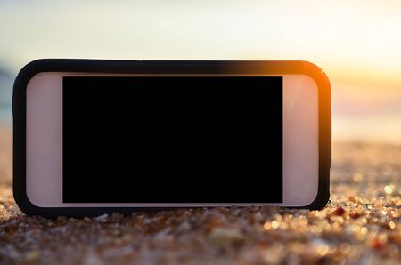 Smart phone on tropical sunset beach with bokeh light sand texture background. Black copy space of technology and travel nature concept. Vintage tone filter effect color style.の写真素材