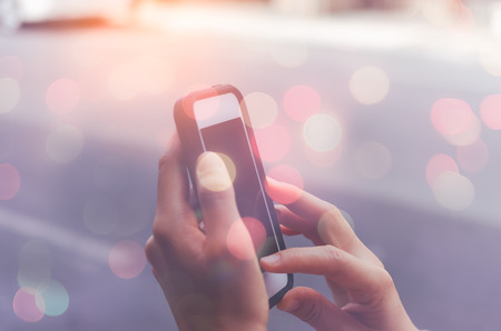 Woman hand using smart phone and blur bokeh sun light on street background. Travel adventure lifestyle and business economic concept. Shallow depth of field. Vintage tone filter effect color style.の写真素材