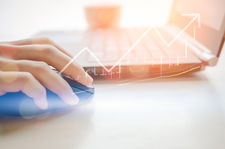 Business economic and technology working concept. Woman hand using mouse notebook double exposure graph money stock trading up trend green arrow bokeh background. Vintage tone filter effect color.の写真素材