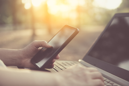Woman hand using smart phone and laptop in outdoor nature park and sunset sky with bokeh light abstract background. Technology business and freelance working concept. Vintage tone filter effect color style.の写真素材