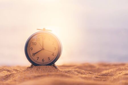 Clock on sand beach in morning suns light with smooth wave background. Copy space of time and summer holiday concept. Vintage tone filter effect color style.の写真素材