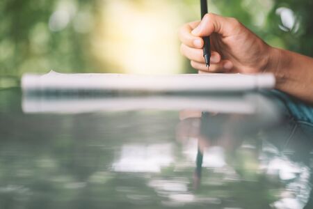 Woman hand using pen writing on paper  at coffee shop with nature green bokeh sun light abstract background. Copy space of education learning and business lifestyle concept. Vintage tone filter color.の写真素材